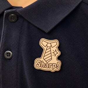 Puede incluir: Un broche de madera clara con forma de corbata con cuello, que presenta rayas diagonales grabadas, un pequeño icono de estrella y la palabra Sharp! en una fuente negrita y redondeada, sujeto a un polo azul marino texturizado.