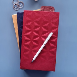 May include: Three notebooks stacked on top of each other. The top notebook is red with a geometric pattern and a white pen lying on top. The middle notebook is navy blue and the bottom notebook is gold.