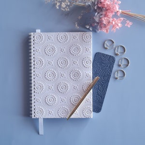 May include: A white, spiral-bound notebook with an embossed floral design, a navy bookmark, and a gold pen are arranged on a light blue surface. Gold paper clips and dried flowers complete the stationery set. The notebook has a matching ribbon.