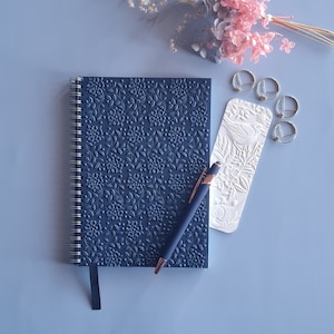 May include: A dark blue notebook with a floral embossed design, a silver spiral binding, and a black ribbon bookmark. A pen and a white bookmark with an embossed floral design are lying on top of the notebook.