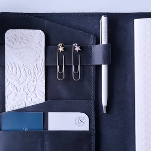 May include: A black leather planner with a white pen, two gold star-shaped paper clips, and a white embossed card with a floral design.