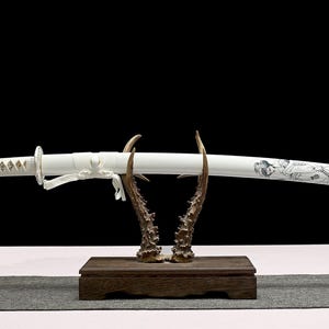 A white katana sword in its white scabbard, featuring a black geisha design, is displayed on a wooden antler stand.