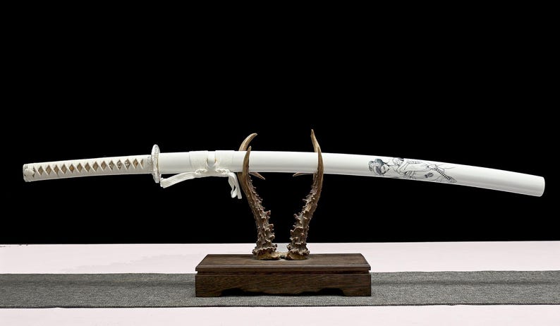 A white katana sword in its white scabbard, featuring a black geisha design, is displayed on a wooden antler stand.
