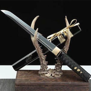 May include: A Japanese tanto knife with a black handle and blade, displayed on a deer antler stand. The knife is in a black scabbard, tied with gold ribbon. The display is set on a wooden base.