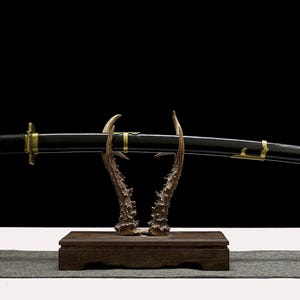 A black and gold katana scabbard with gold accents rests on a wooden stand with antler-like supports.