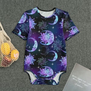 May include: A short-sleeved baby bodysuit featuring a celestial design. The bodysuit has a black background with a pattern of purple and teal suns, moons, and stars. The garment is made of a soft material and has a white interior.