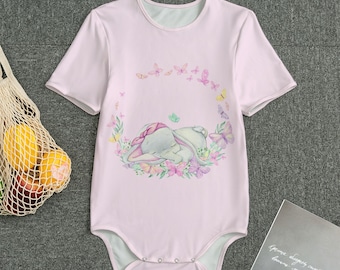 Könnte beinhalten: Ein hellrosa Baby-Body mit kurzen Ärmeln. Er zeigt eine Aquarellillustration eines schlafenden Hasen, umgeben von einem Kranz aus Blumen und Schmetterlingen. Der Hintergrund ist einfarbig hellrosa.