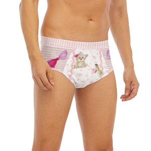Puede incluir: Ropa interior blanca y rosa con un diseño de oso de peluche y lazo. La ropa interior presenta un patrón de rayas rosas y blancas en los laterales y un panel frontal blanco con un oso de peluche, corazones y otros gráficos. El oso de peluche tiene un lazo rosa.