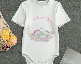 Könnte beinhalten: Weißer Baby-Body mit kurzen Ärmeln. Er zeigt eine Aquarell-Illustration eines schlafenden Hasen, umgeben von einem Kranz aus Blumen und Schmetterlingen in Rosa, Lila und Gelb. Der Body hat Druckknöpfe.
