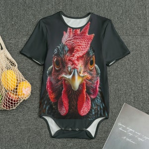 May include: Black baby bodysuit featuring a close-up of a rooster's face. The rooster has red comb and wattle, with detailed feathers and a focused gaze. The bodysuit has short sleeves and a snap closure.