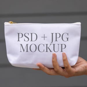May include: A white canvas cosmetic bag with a gold zip, held in a person's hand. The bag features the text "PSD + JPG MOCKUP" in grey, centered on the front. The background is a muted grey.