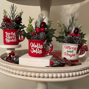 May include: Three red mugs with white holiday-themed text and green and red floral arrangements. The mugs read "Joyful Merry Blessed", "Holly Jolly", and "Merry and Bright". The mugs are on a white tiered serving tray.