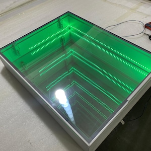 Może przedstawiać: Białe kwadratowe pudełko z szklanym wierzchem. Szkło jest lustrzane i ma zielone diody LED wokół obwodu. Diody LED tworzą efekt odbicia.