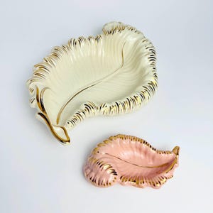 Puede incluir: Dos platos de cerámica con forma de hoja. Uno es blanquecino con ribete dorado y detalle de veta dorada. El otro es rosa con ribete dorado. Ambos tienen un acabado brillante.