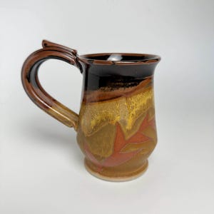 May include: A brown ceramic mug with a unique handle and a glaze that features shades of brown, yellow, and orange. The mug has a geometric design with a leaf-like pattern.