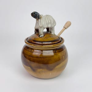 May include: A brown ceramic jar with a lid featuring a turtle figurine and a wooden spoon. The jar has a round shape with a glossy finish. The turtle is white and black. The spoon is light brown.