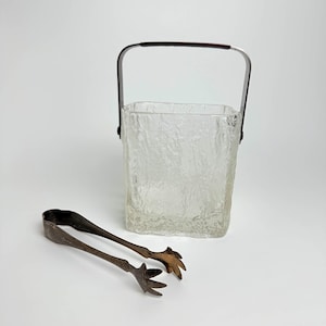 Könnte beinhalten: Ein durchsichtiger Eiseimer aus Glas mit strukturierter Oberfläche und einem silbernen Griff. Zum Eimer gehört eine silberne Eis-Zange mit einem klauenartigen Design.