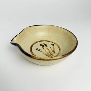 May include: A beige ceramic bowl with a spout and a brown rim. The bowl features a circular design with a brown floral pattern in the center. The bowl is likely used for serving sauces or dips.