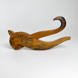 Op de afbeelding: Een houten notenkraker gegraveerd in de vorm van een vis met een gedetailleerd bloemenontwerp. De notenkraker is bruin en heeft een gebogen handvat.