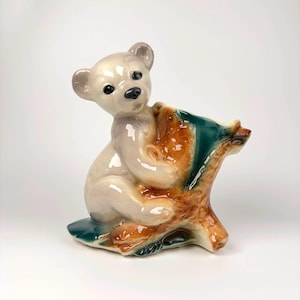 Pode incluir: Estatueta de cerâmica de um urso de pelúcia abraçando um tronco de árvore. O urso é branco sujo com olhos e nariz pretos. O tronco é marrom e verde. Um item decorativo.