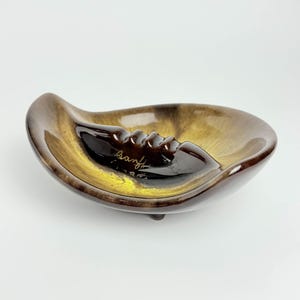 May include: A brown and yellow ceramic ashtray with three legs. The ashtray has a wave-like shape and says "Banff" in gold lettering.
