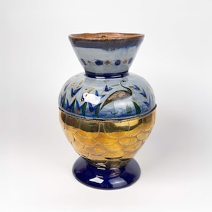 May include: A decorative vase in blue, gold, and brown. The top section is blue with a brown rim and a painted design. The middle section is gold with a scale-like pattern, and the base is a dark blue. The vase is approximately 30 cm tall.