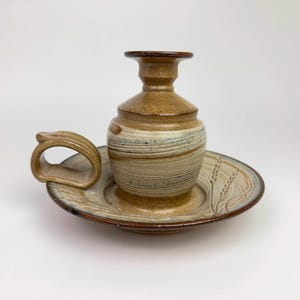 May include: A handcrafted ceramic candle holder set. The set includes a small, brown and beige vase-shaped candle holder, a matching plate with wheat stalk detailing, and a handle. The pottery has a rustic, earthy aesthetic.