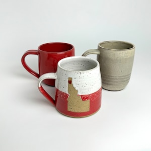 May include: Three ceramic mugs, one red, one white with brown speckles and a brown Idaho state outline, and one light gray. The mugs are all handmade and have a rustic look.