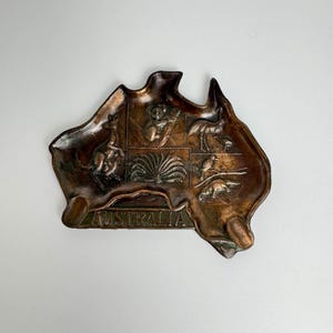 May include: A copper-colored metal ashtray shaped like the continent of Australia. The ashtray features embossed images of native Australian animals, including a kangaroo, koala, emu, and a bird. The word "AUSTRALIA" is embossed on the bottom of the ashtray.