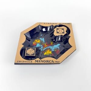 Puede incluir: Un cenicero de cerámica con interior azul oscuro y borde marrón. El cenicero presenta un diseño abstracto colorido con detalles en amarillo, rojo y azul. El texto "ceramica MINORCA ITALY" está impreso en la parte delantera.