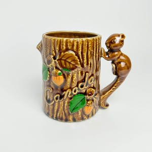 May include: A brown ceramic mug shaped like a tree trunk with a squirrel handle. The mug has the word "Canada" written on it and features acorns and leaves.