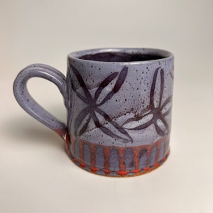 May include: A purple ceramic mug with a black floral design and a red and orange striped band around the bottom.
