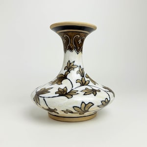Può includere: Un vaso decorativo in ceramica con base bianca e disegno floreale marrone. Il vaso ha un corpo bulboso e un collo svasato con accenti marroni e beige. Il motivo floreale è nelle tonalità del marrone, il vaso è alto circa 15 cm.