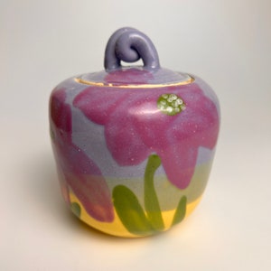 Könnte beinhalten: Eine kleine Keramikdose mit Deckel, handbemalt mit lila und grünen Blumen auf gelbem Hintergrund.