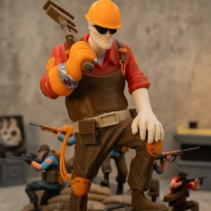 May include: A figurine of an engineer in an orange hard hat and sunglasses, holding a wrench. The engineer stands on a red toolbox with the number 24. Other figurines are in the background, along with tools and ammunition.