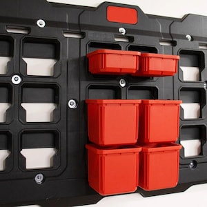 May include: A black wall-mounted storage system with multiple compartments and red storage bins. The system features a grid-like design with various sized openings for organization. The red bins are rectangular and designed to fit into the system's openings.