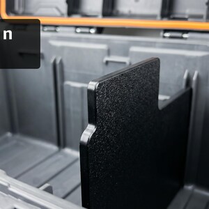 以下が含まれることがあります： 黒い仕切りが付いた濃い灰色の収納容器の内部ビュー。「Drop In Fit」というテキストが黒い長方形に表示されています。容器にはオレンジ色のアクセントがあります。