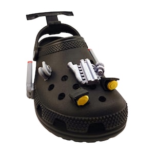 Puede incluir: Zueco negro adornado con abalorios con temática automovilística. El zapato presenta una base negra con varios detalles grises y amarillos, incluyendo un turbocompresor, tubos de escape y faros. El zapato tiene una correa de talón pivotante.