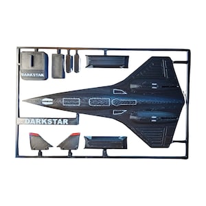 May include: A black plastic model kit of a Darkstar aircraft. The kit includes the main body, wings, and other components, all still attached to the sprue. The word "DARKSTAR" is printed on the sprue.