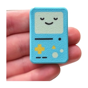 May include: A light blue, rectangular pin featuring a smiling face and a retro video game design. The pin has a textured surface and a yellow cross-shaped button. The design is reminiscent of a classic handheld gaming device.