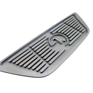 Puede incluir: Una alfombrilla de piso de coche de goma gris con un logotipo en relieve de una letra estilizada "G" dentro de un c&iacute;rculo. La alfombrilla tiene una superficie texturizada con l&iacute;neas en relieve.
