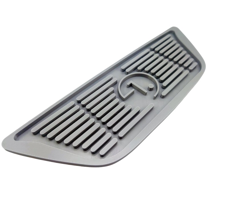 Puede incluir: Una alfombrilla de piso de coche de goma gris con un logotipo en relieve de una letra estilizada "G" dentro de un c&iacute;rculo. La alfombrilla tiene una superficie texturizada con l&iacute;neas en relieve.