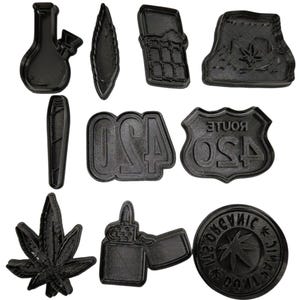 Puede incluir: Un conjunto de cortadores de galletas negros en varias formas, incluyendo un bong, una hoja, un teléfono móvil, una señal de tráfico con "420", una hoja de marihuana, un encendedor y un emblema redondo con texto y hoja.