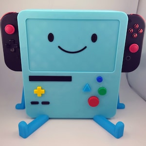 Puede incluir: Una consola Nintendo Switch azul y negra con un soporte, que presenta una cara sonriente en la pantalla. La consola tiene un botón amarillo, rojo, verde y azul en la parte delantera.