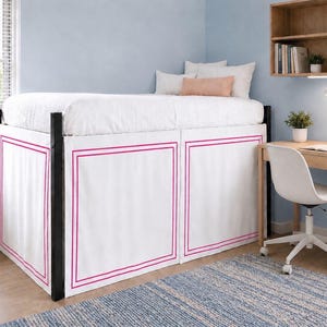 Könnte beinhalten: Ein weißer Bettvolant mit einem doppelten rosa Rahmenmuster, der um ein erhöhtes Bettgestell mit schwarzen Metallpfosten angebracht ist. Das Bett ist mit einer weißen Bettdecke und mehreren Kissen ausgestattet, darunter ein pfirsichfarbenes Kissen.