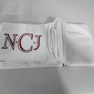 May include: White bedding set with embroidered burgundy initials "NCJ". The set is neatly folded, displaying the monogram on the top sheet. The bedding appears to be made of a smooth, crisp fabric.