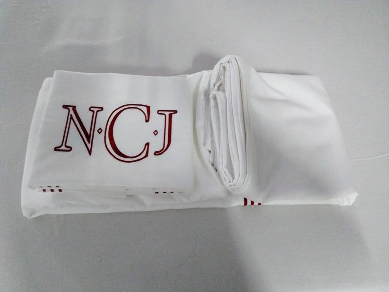 May include: White bedding set with embroidered burgundy initials "NCJ". The set is neatly folded, displaying the monogram on the top sheet. The bedding appears to be made of a smooth, crisp fabric.