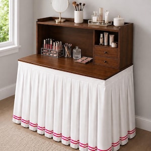 May include: A dark wood vanity desk featuring a hutch with two small drawers and open shelving. The base is covered by a white pleated fabric skirt with two horizontal pink stripes along the bottom edge, holding various makeup and beauty products.