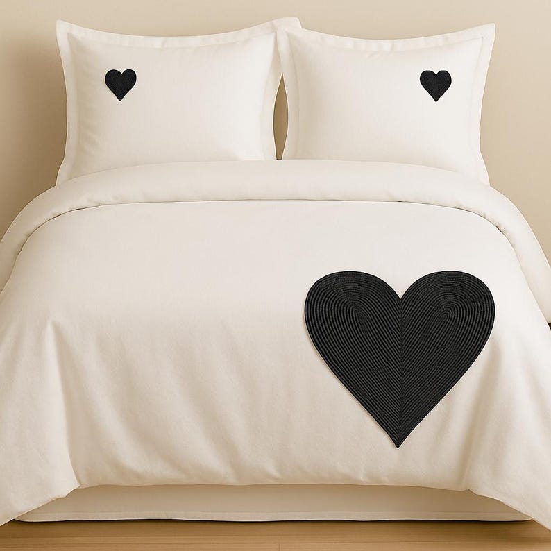 May include: A cream-colored bedding set consisting of a duvet cover and two pillowcases. Each pillowcase features a small, solid black embroidered heart. The duvet cover displays a large, textured black heart with a ribbed, concentric pattern.