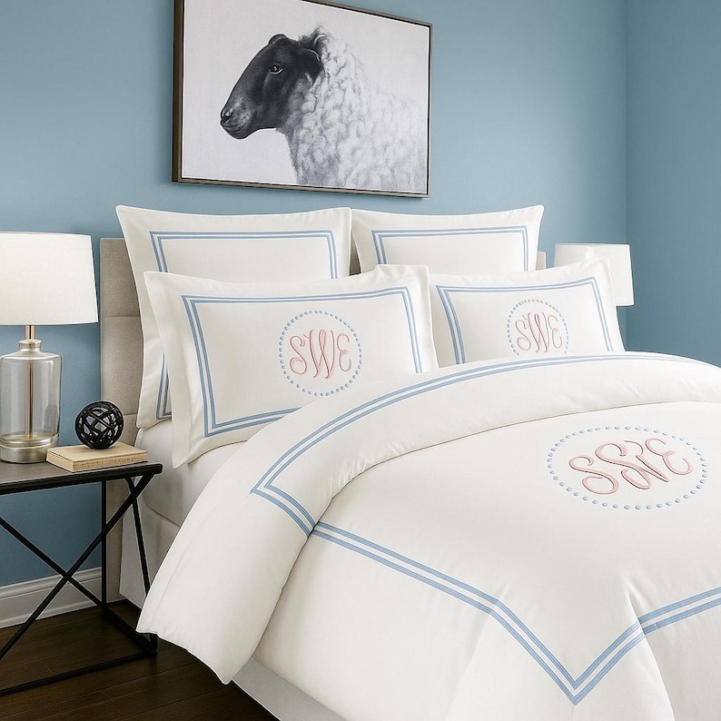May include: White bedding set with light blue trim and embroidered initials. The set includes a duvet cover and pillowcases. A framed black and white sheep portrait hangs on the blue wall above the bed. A lamp and side table are visible.